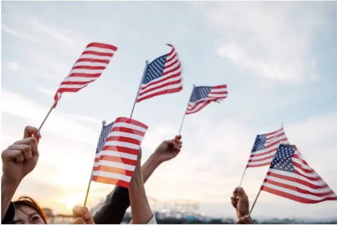 Several hands raise small American flags against a bright sky.